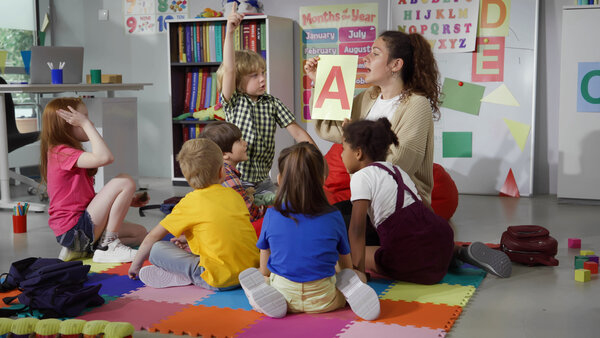 Bild vergr&ouml;&szlig;ern: Eine Person sitzt mit Vorschulkindern im Kreis und zeigt diesen Buchstaben